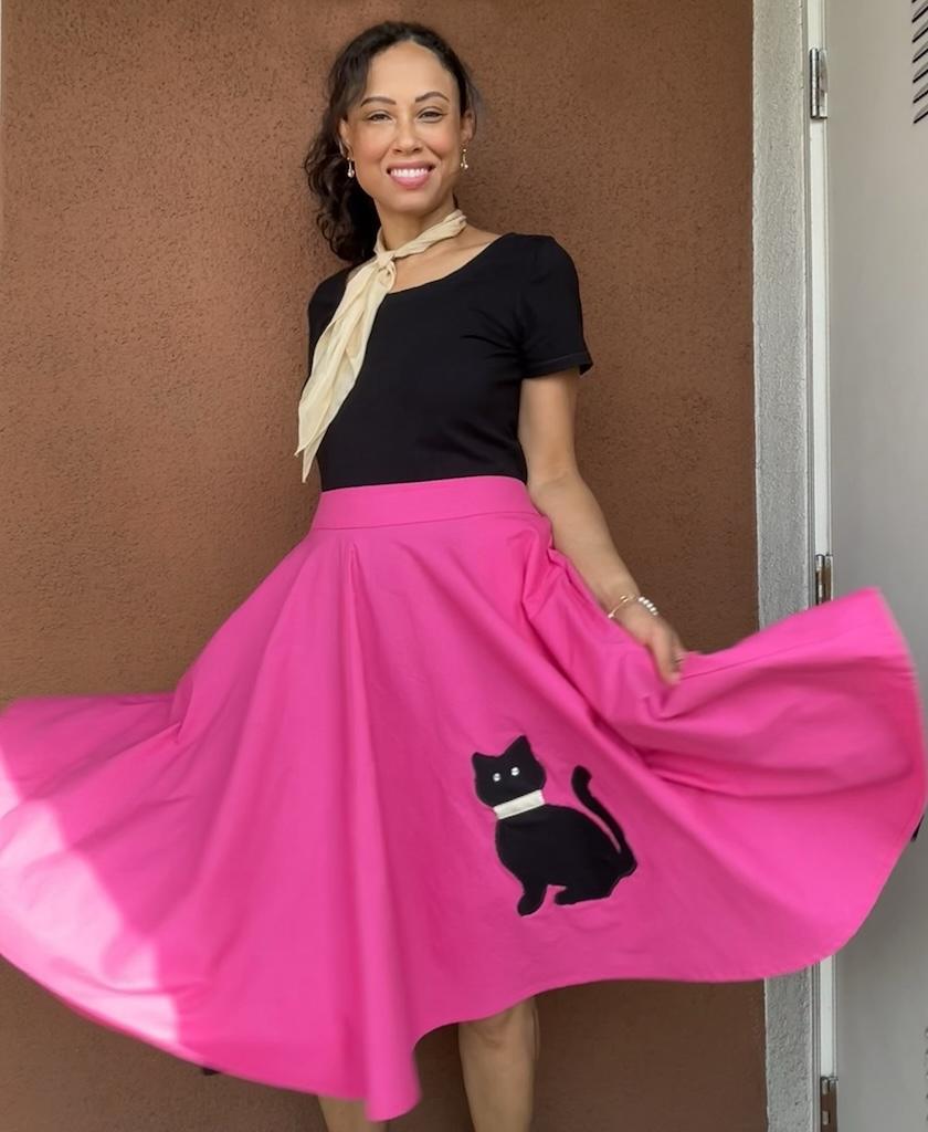 Luna twirling in a bright pink sock hop skirt with black cat applique in the front, black shirt, and beige scarf