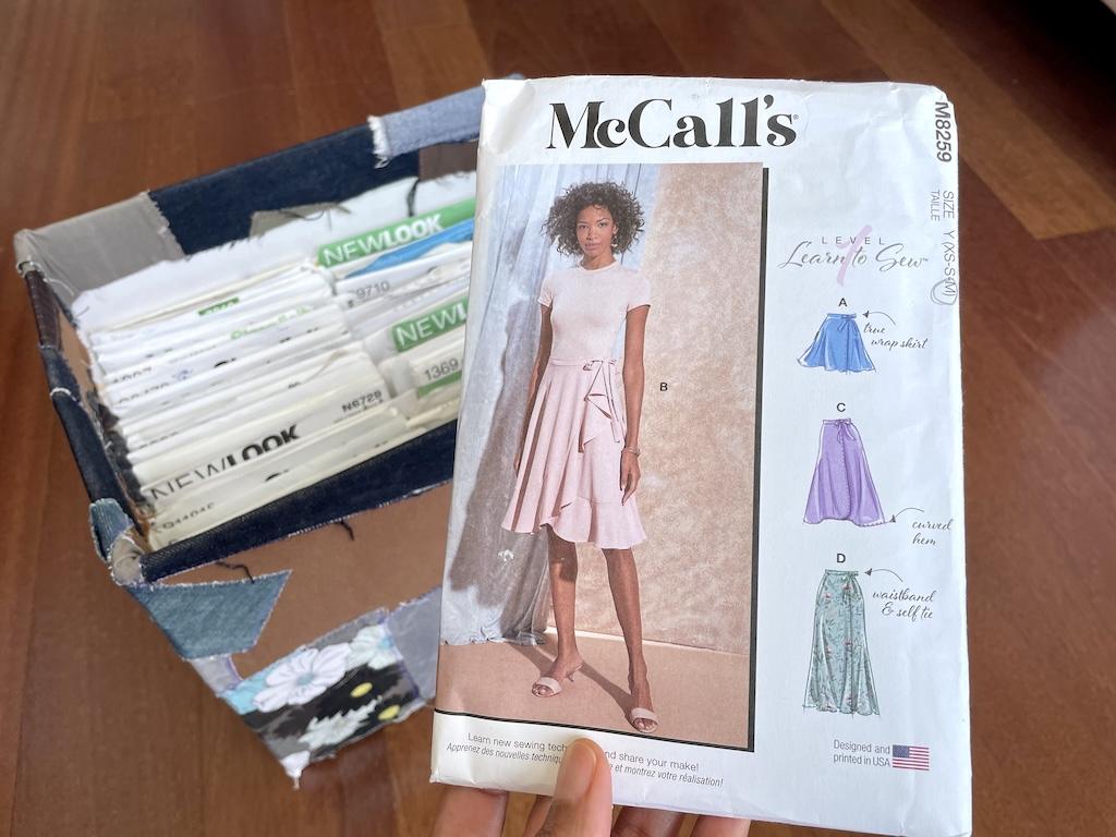 Luna’s hand holding up McCall’s learn to sew skirt pattern with her box of sewing patterns in the background