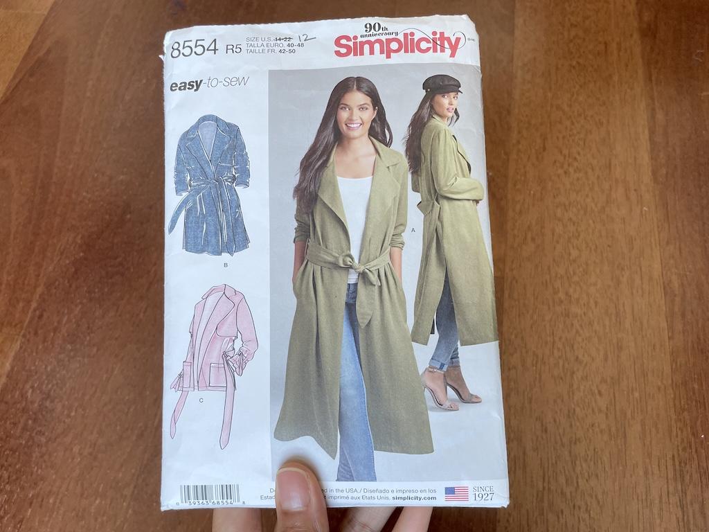 Luna’s hand holding Simplicity S8554 sewing pattern envelope with model wearing soft trench coat