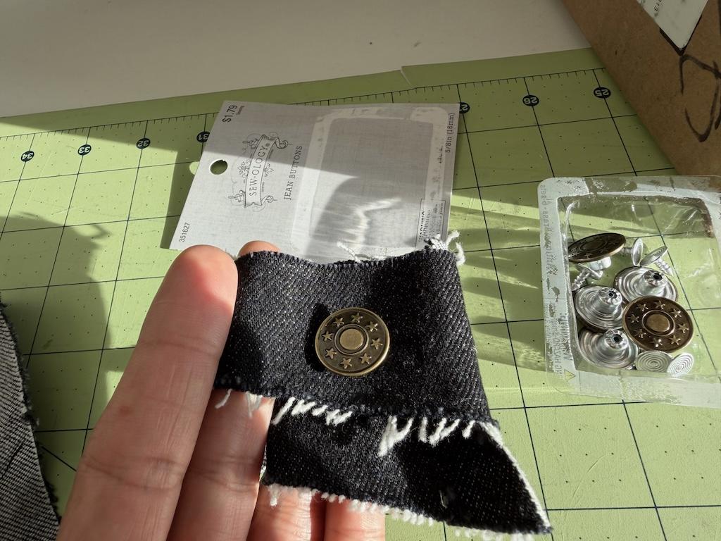 Luna’s fingers holding scrap fabric with sewology jeans button attached with package in background