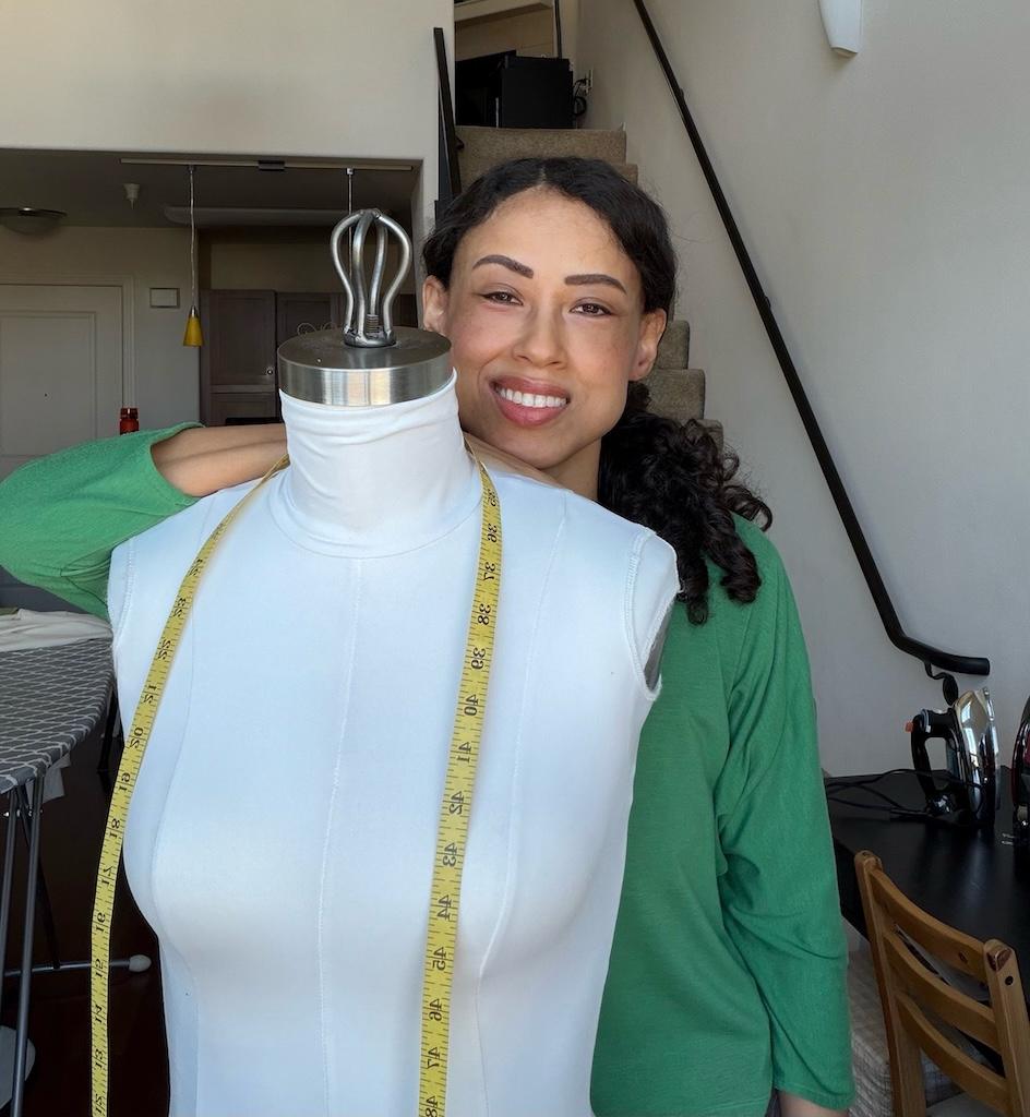 Luna standing behind dress form and smiling to camera in her sewing room